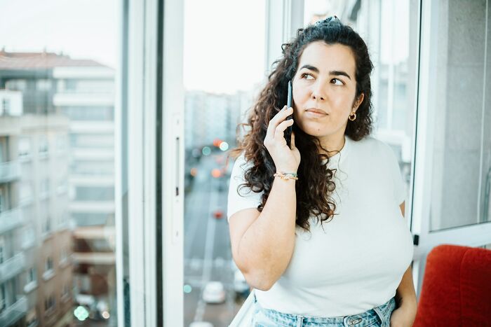 Woman with curly hair wearing a white shirt, talking on phone near window with city view, representing boomer opinions.