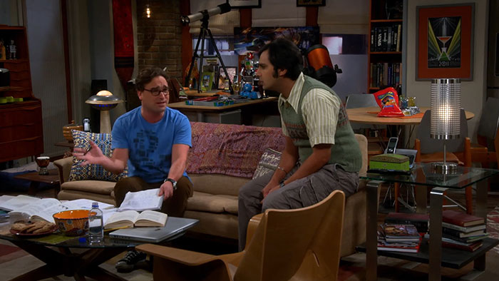 Two men having a conversation in a living room filled with books and snacks, relating to celebrities and hygiene habits.