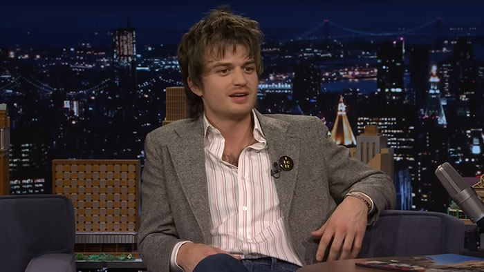 Young male celebrity sitting on a late-night talk show set, discussing hygiene habits amid city night skyline backdrop.