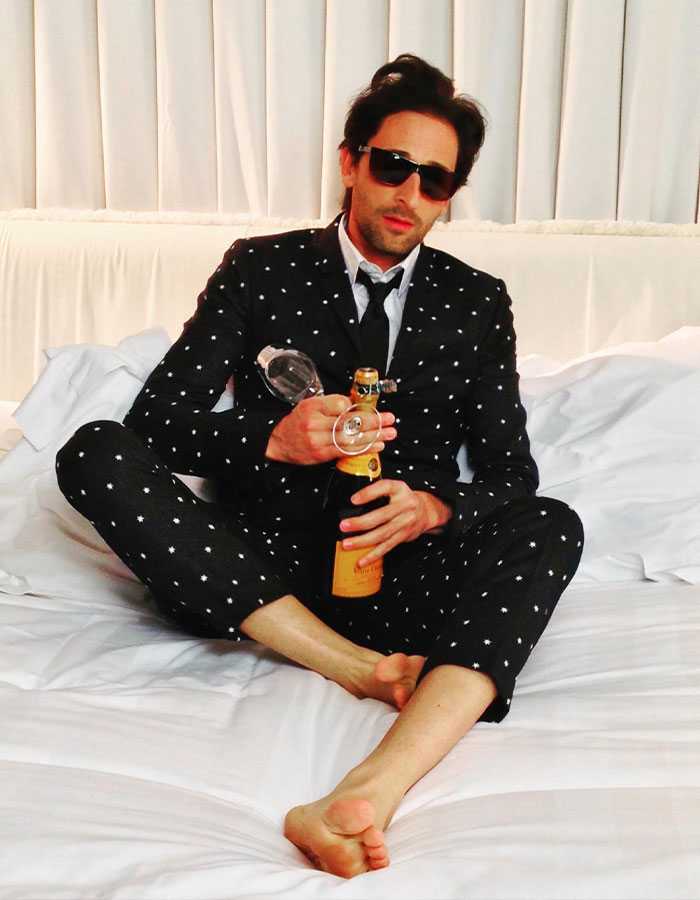 Adrien Brody sitting on a bed in a patterned suit, holding a champagne bottle and glass, wearing sunglasses. Adrien Brody sitting on a bed in a patterned suit, holding a champagne bottle and glass, wearing sunglasses.