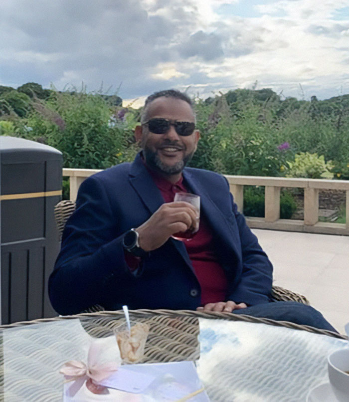 Man in sunglasses and navy blazer sitting outdoors holding a drink, related to father of four with 37 followers jailed over tweet.