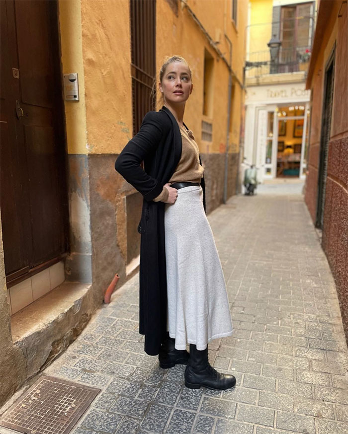 Amber Heard standing in a narrow street wearing a long black coat and light skirt, illustrating net worth timeline context.