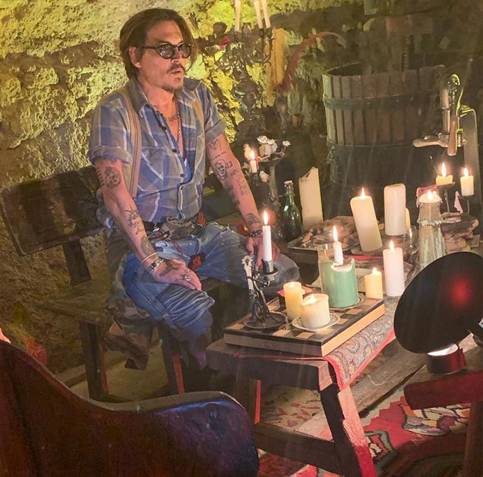 Johnny Depp with tattoos sitting near a table full of lit candles in a dimly lit rustic setting discussing Amber Heard net worth timeline.