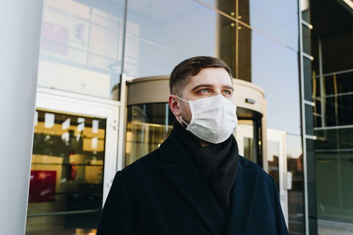 A man wearing a white face mask and dark coat standing outside a modern building, illustrating life hacks concept.