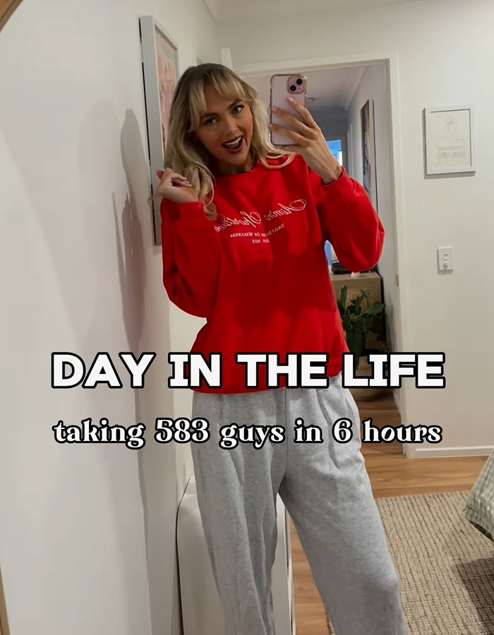 Adult content creator taking selfie wearing red sweatshirt and gray pants in hallway with text about 583 men in 6 hours.
