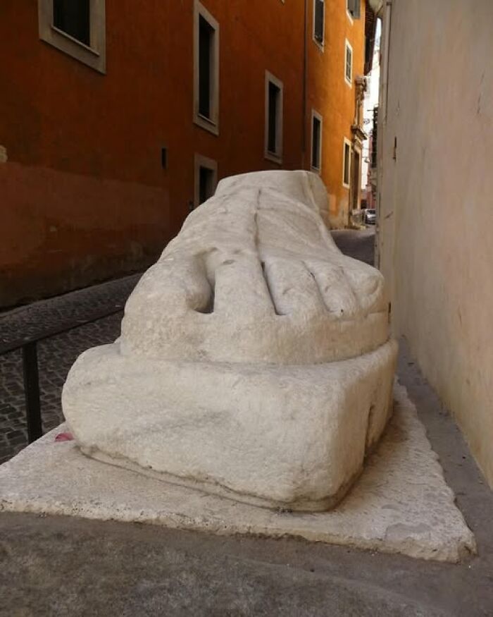 Large ancient stone sculpture of a foot displayed outdoors in a narrow street representing archeology and art.
