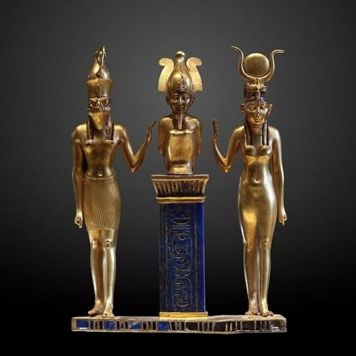 Golden archeological art statue depicting three ancient Egyptian deities on a detailed blue pedestal against a dark background.