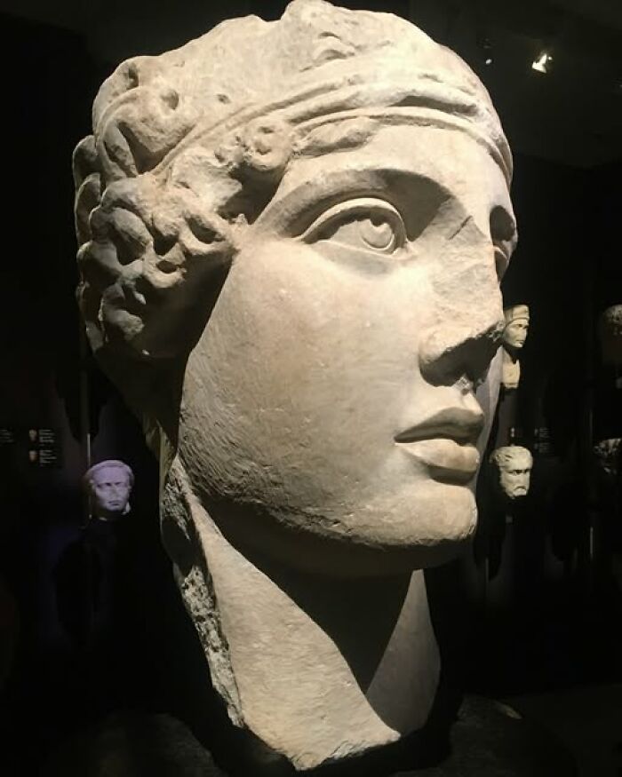 Large ancient stone sculpture of a human head displayed in a dim museum setting showcasing archeology and art.