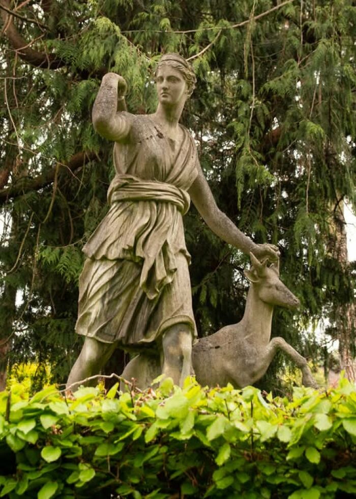 Statue of a woman and deer surrounded by greenery, representing archeology and art in an outdoor natural setting.