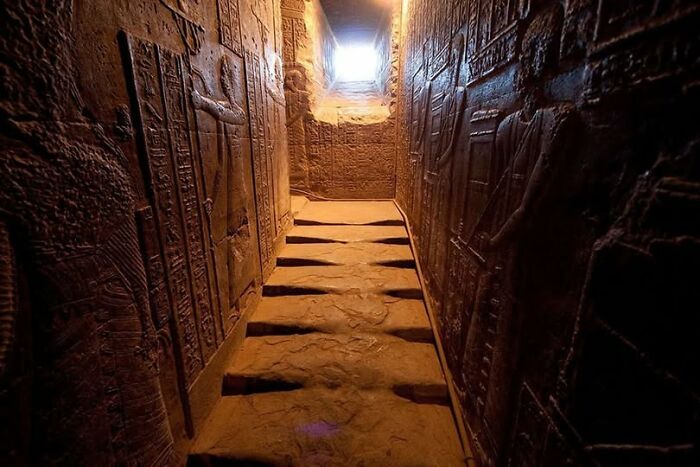 Ancient archeological corridor with detailed wall carvings and art illuminated by light from an opening above.