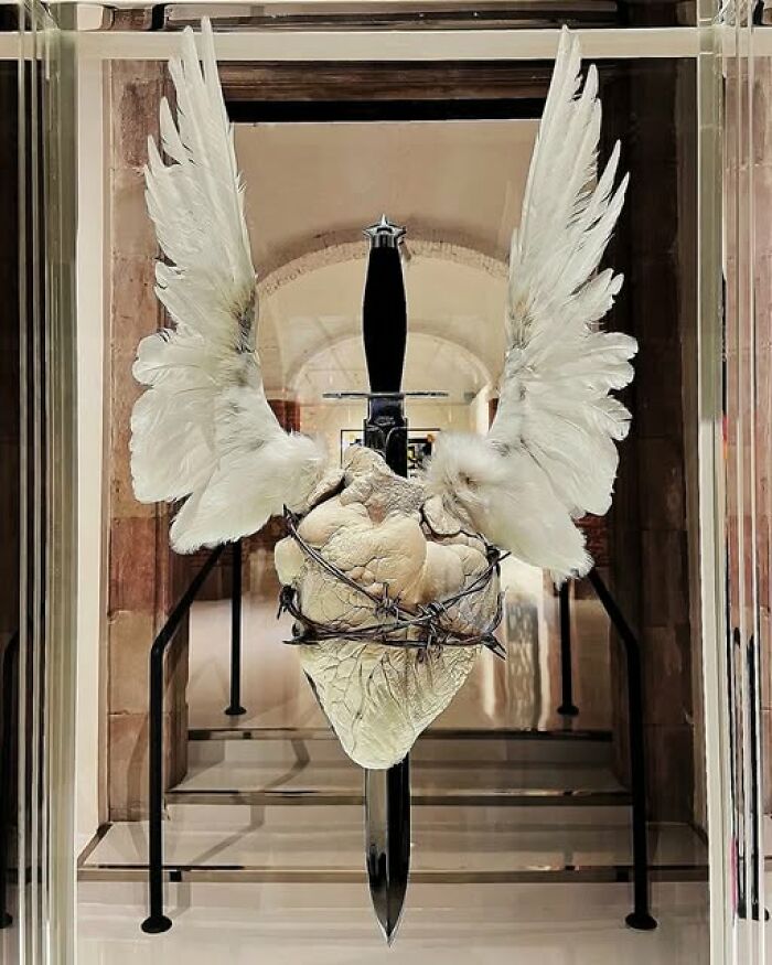 Sculpture of a heart wrapped in barbed wire with white wings and a sword behind it in an archeology and art exhibit.