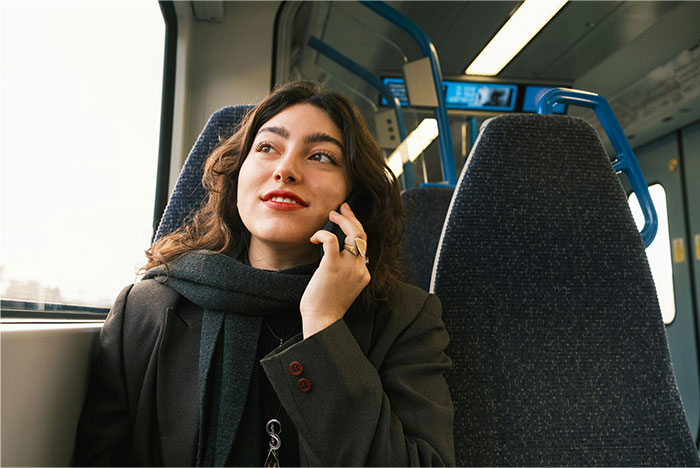 Young woman on public transport, speaking on phone, considering asking roommates to leave with thoughtful expression.