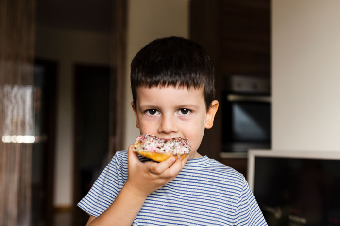 Young boy eating a doughnut in a home kitchen, illustrating food talk triggers related to son’s autism challenges. Young boy eating a doughnut in a home kitchen, illustrating food talk triggers related to son’s autism challenges.