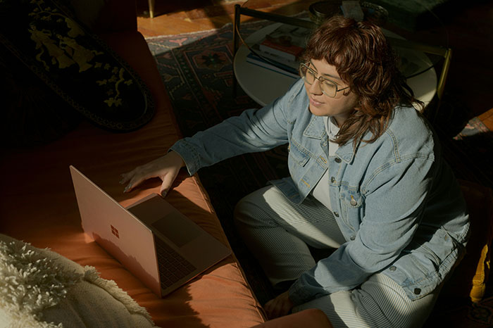 Person in a denim jacket sitting on the floor, using a laptop in a cozy room with warm natural light.