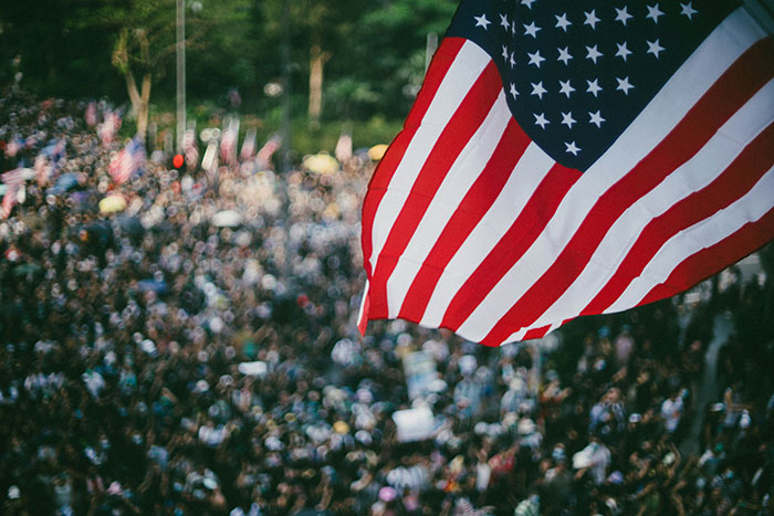 American flag waving above a large crowd, symbolizing the biggest elephant or blue whale in the room.