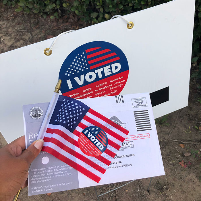 Hand holding American flag, ballot envelope with I voted sticker, outside near a yard sign in grassy area.