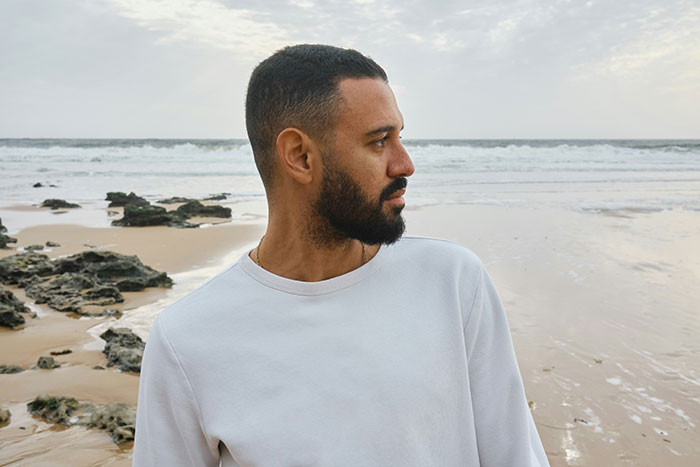 Man with beard wearing white sweater looking at ocean waves on a rocky beach with overcast sky, blue whale in the room concept.