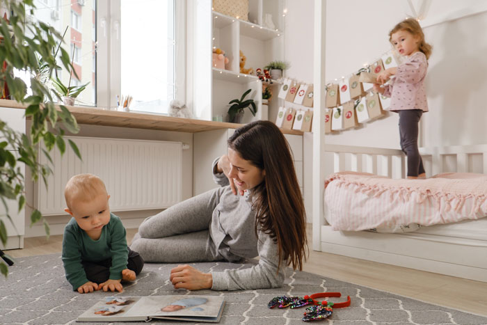 18-year-old woman caring for two kids in a bright room, playing with them while lying on a carpeted floor.