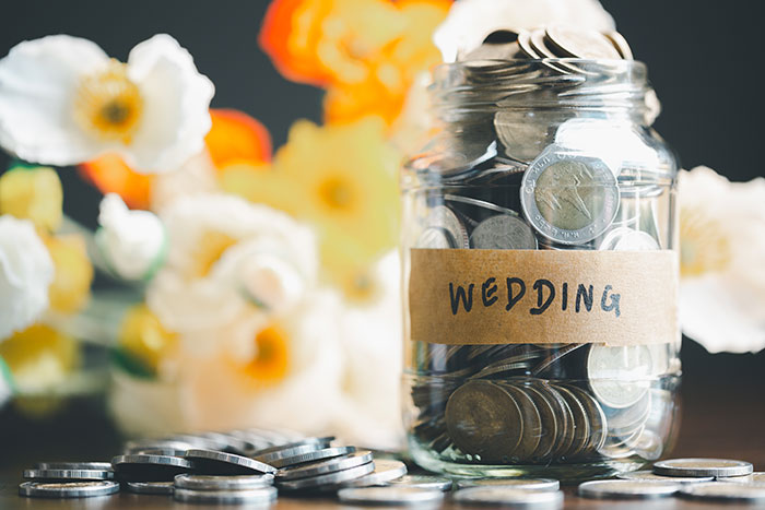 Glass jar labeled wedding filled with coins surrounded by flowers, relating to paying for hen parties debates online.