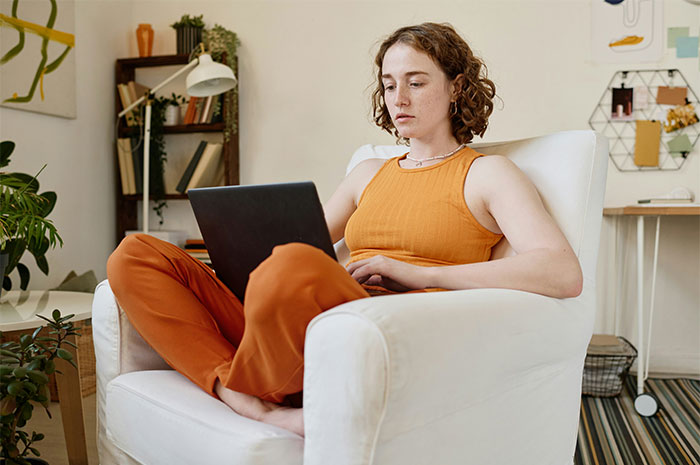 Young woman in orange outfit sitting in armchair using laptop, related to HR employees sharing worst background checks.