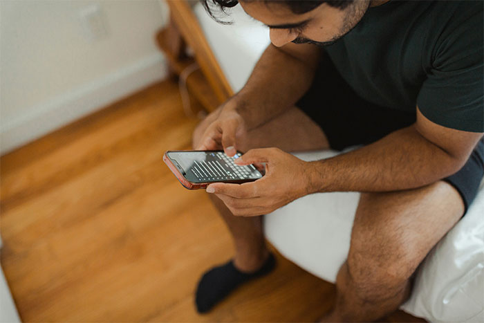 Man in casual clothing checking phone messages while sitting on bed, illustrating HR employees reviewing background checks.