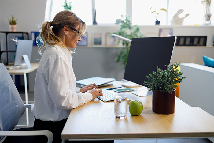 HR employee wearing glasses typing on keyboard at desk with plant, apple, and glass of water in bright office setting.