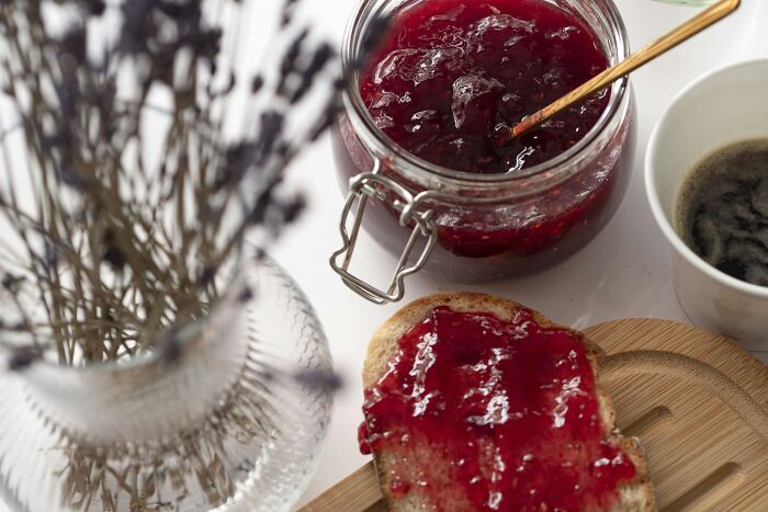 Jar of red jam with wooden spoon, jam spread on bread, cup of coffee, and dried flowers in a glass vase.