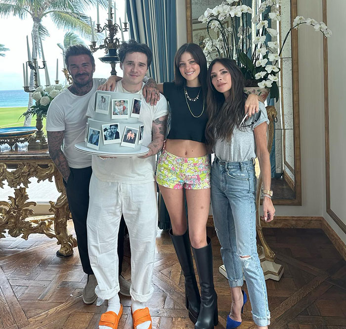 David and Brooklyn Beckham with Nicola Peltz and Victoria Beckham posing indoors with a decorated cake near a window.