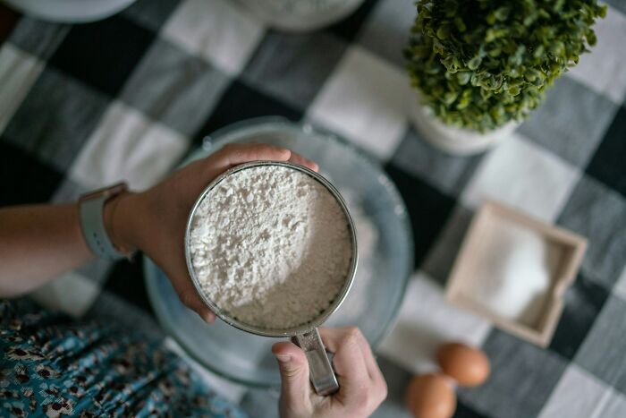 Person holding a measuring cup of flour preparing ingredients for baking in a food prices comparison context