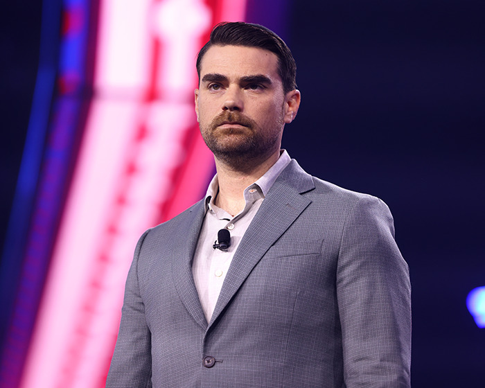 Ben Shapiro in a gray suit speaking at an event, facing forward with a serious expression on stage. Ben Shapiro in a gray suit speaking at an event, facing forward with a serious expression on stage.
