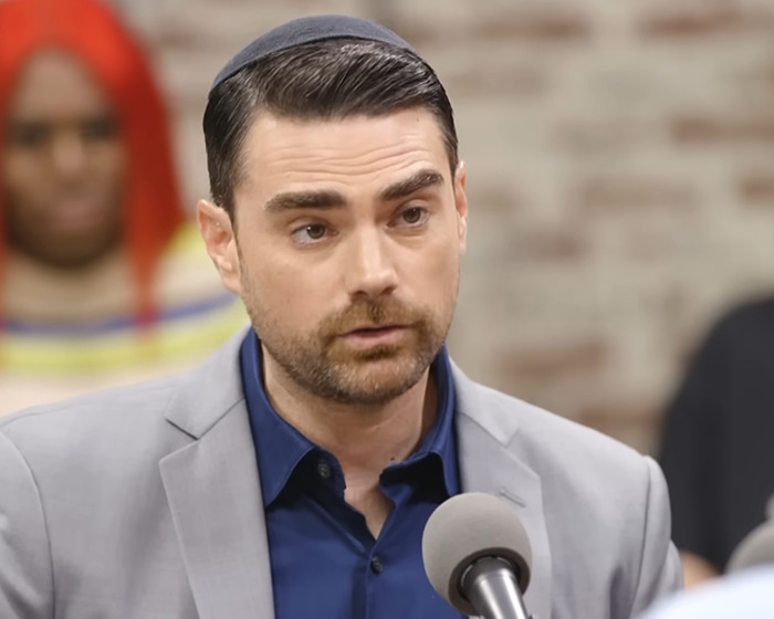 Ben Shapiro speaking into a microphone during a discussion, with blurred people and brick wall in the background. Ben Shapiro speaking into a microphone during a discussion, with blurred people and brick wall in the background.