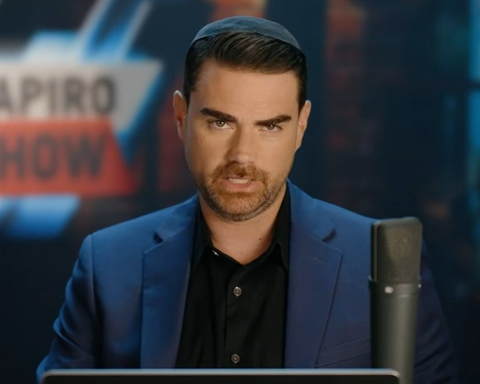 Ben Shapiro speaking on his show wearing a blue blazer and black shirt with a microphone in front of him. Ben Shapiro speaking on his show wearing a blue blazer and black shirt with a microphone in front of him.