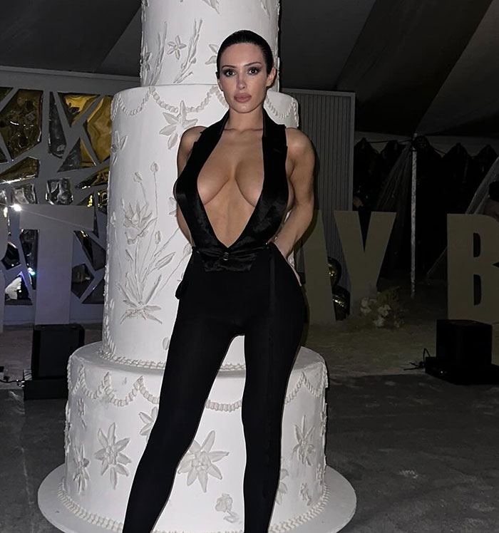 Bianca Censori posing in a revealing black outfit with a deep neckline at a nighttime event setting.