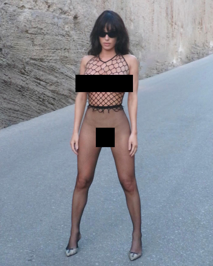 Bianca Censori posing outdoors in revealing fishnet outfit with sunglasses, sparking public calls for arrest over indecency.