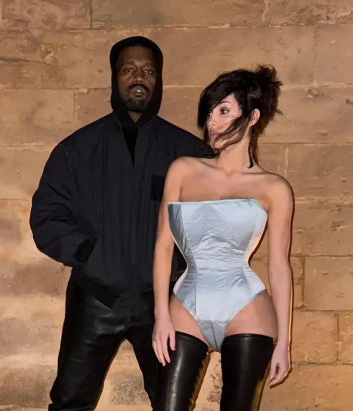 Bianca Censori shows off her butt in a racy outfit with thigh-high boots standing next to a man in a dark hoodie.