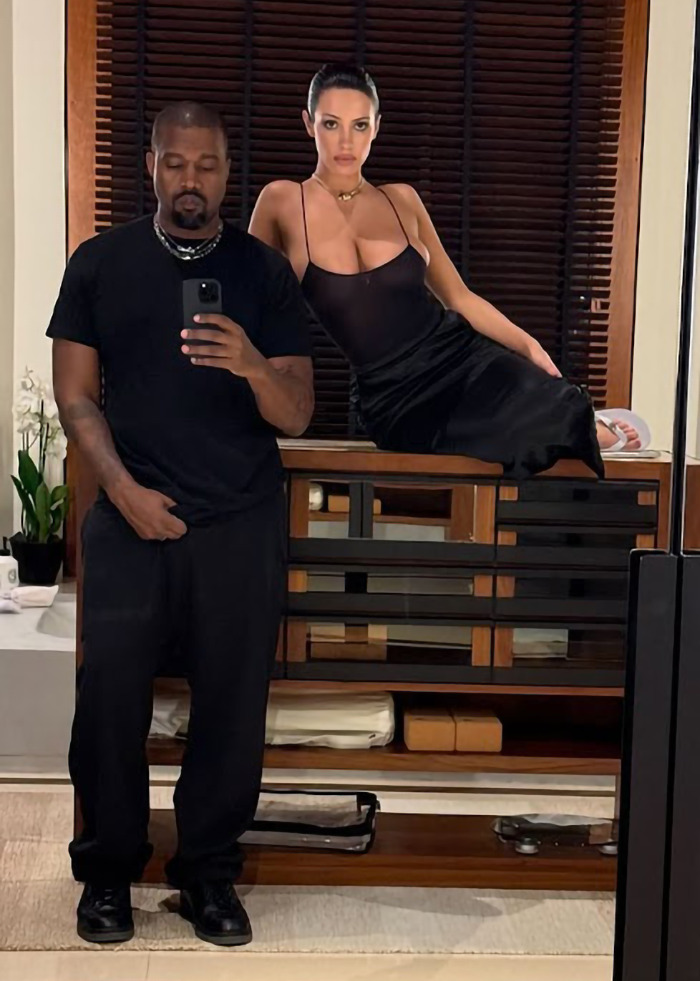 Bianca Censori poses in a sheer top with black skirt and man in black t-shirt taking a mirror selfie indoors.