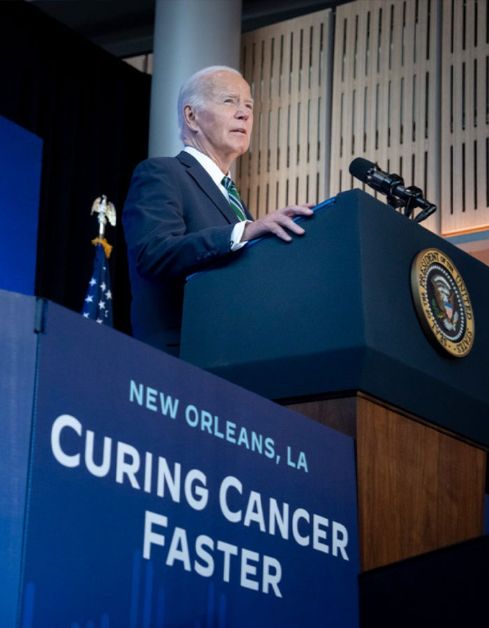 Former President Biden speaking at a podium in New Orleans, emphasizing aggressive prostate cancer and bone spread awareness.