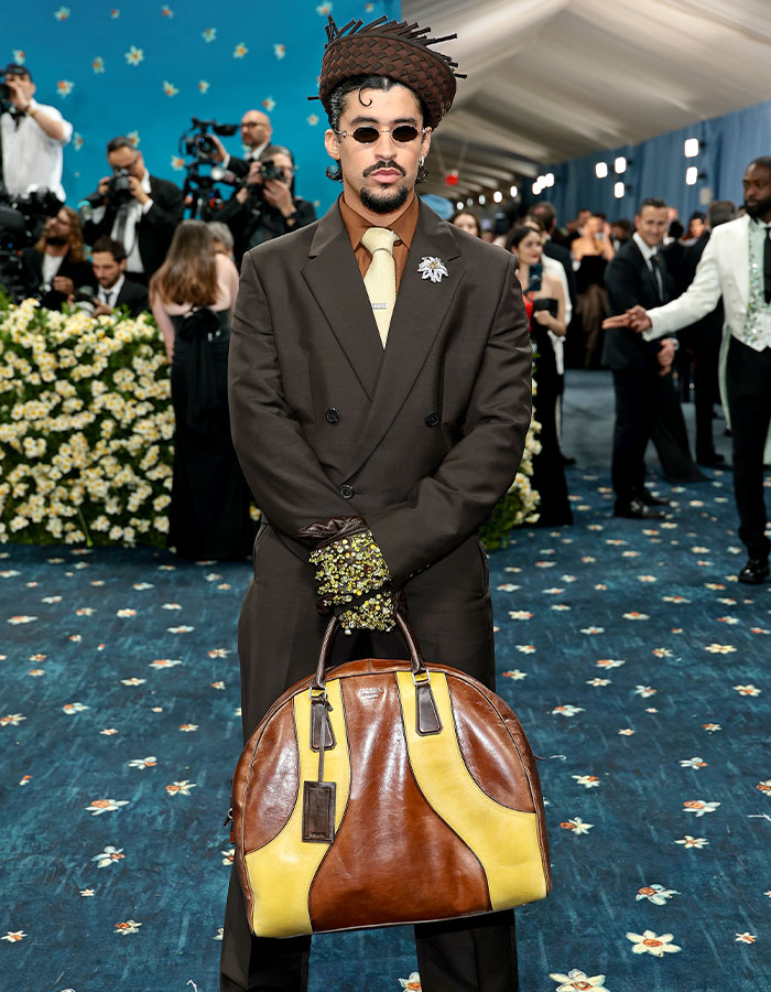 Man wearing oversized hat and gloves holding a large brown and yellow bag at a fashion event, showcasing Met Gala fashion fails.