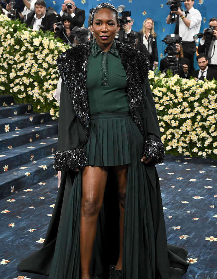 Woman in dark green pleated dress and embellished coat posing on a floral carpet at Met Gala fashion event 2025.