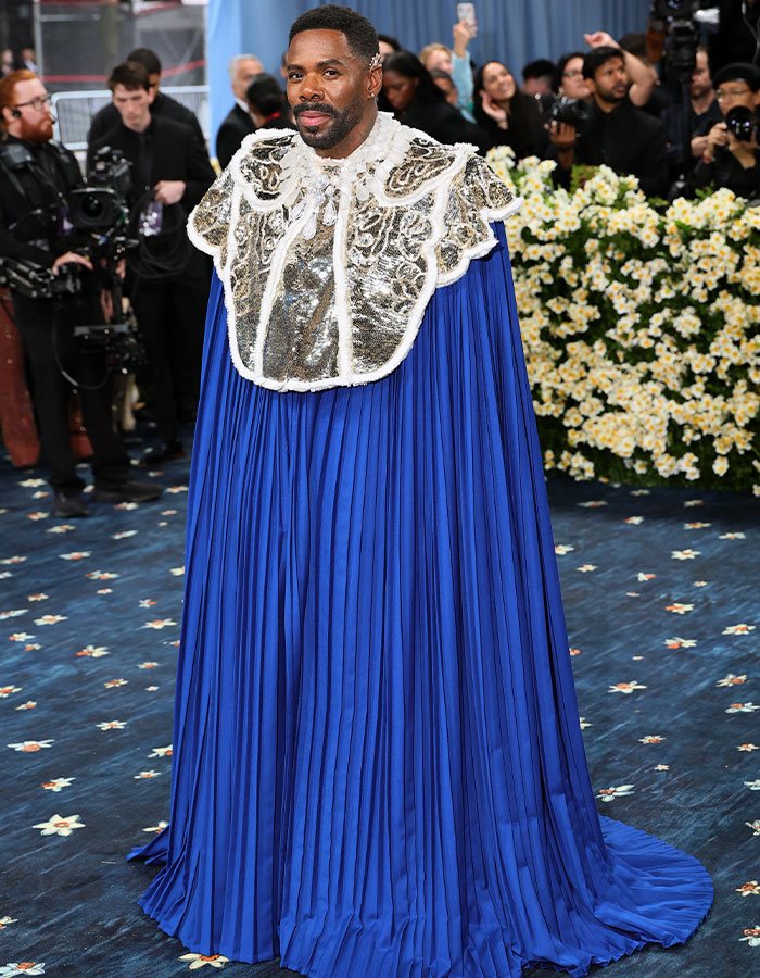 Man wearing an ornate blue and silver cape among photographers and floral decorations at a fashion event with fashion fails theme.