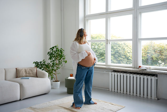 Pregnant woman standing near window, wearing headphones and casual clothes, anticipating her upcoming delivery experience.