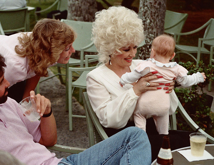 Woman with curly blonde hair holding a baby while people enjoy drinks outdoors in a casual celeb encounter setting.