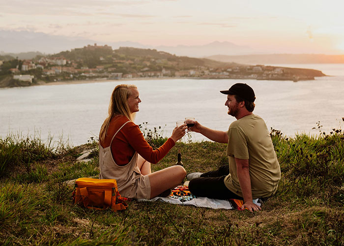 Couple enjoying a scenic outdoor date picnic by the water during sunset, hinting at guy always bringing BFF to dates.