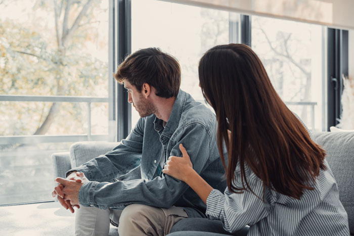 Couple in open relationship having a tense conversation on couch, woman comforting upset man looking away by window.