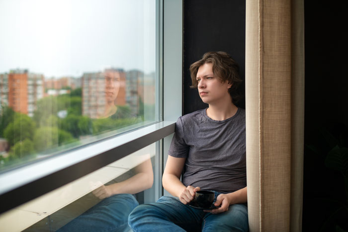 Young man sitting by the window, reflecting thoughtfully, illustrating the impact of helicopter parents on relationships.