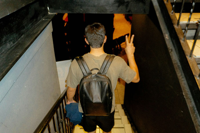 Young guy with backpack walking down stairs, making a peace sign, showing displeasure with girlfriend's school song performance connection.