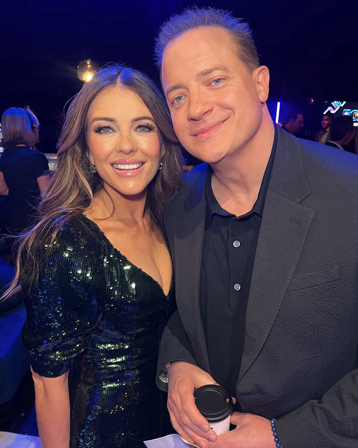 Brendan Fraser with a woman at a cancer gala, showcasing his slimmed down physique and smiling in a dark suit and sequined dress.