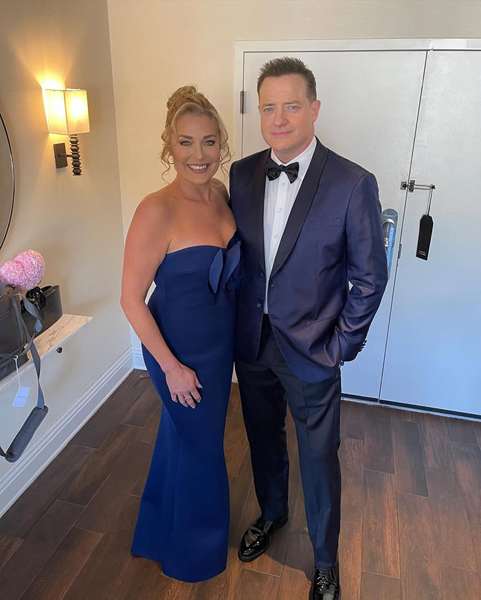 Brendan Fraser in a slimmed down physique wearing a tuxedo, standing next to a woman in a navy blue gown indoors.