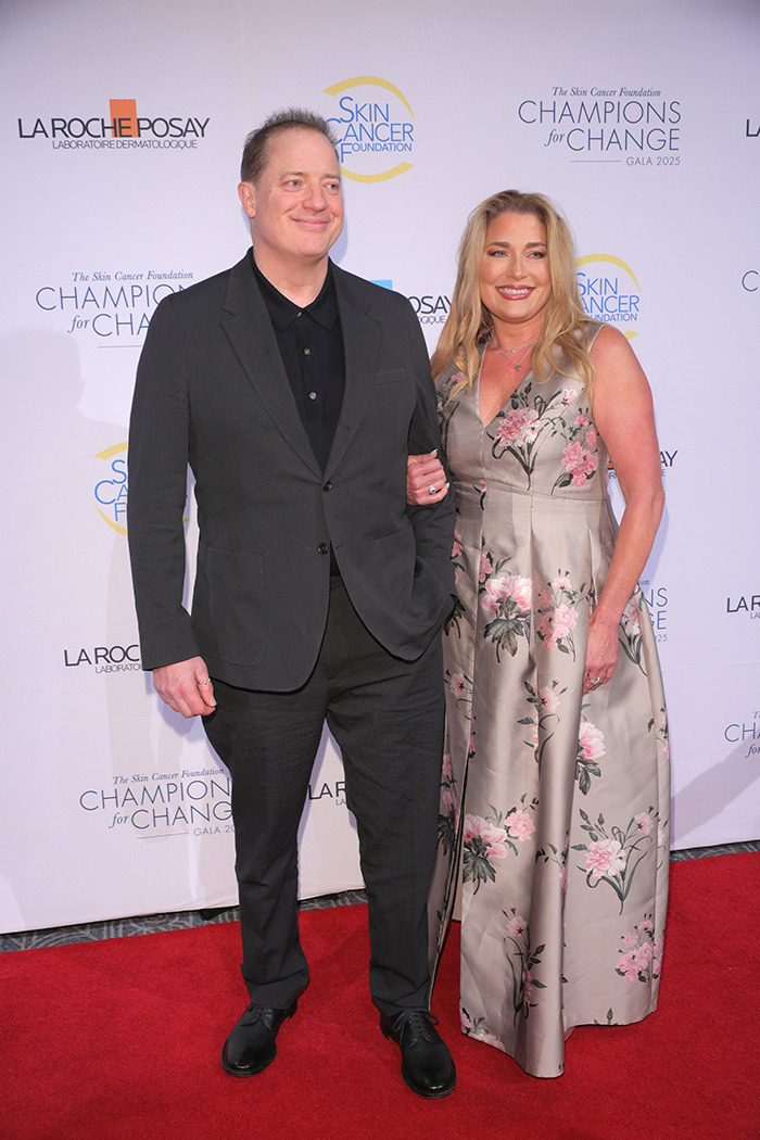 Brendan Fraser in a black suit with a woman in a floral dress on the red carpet at a skin cancer gala event.