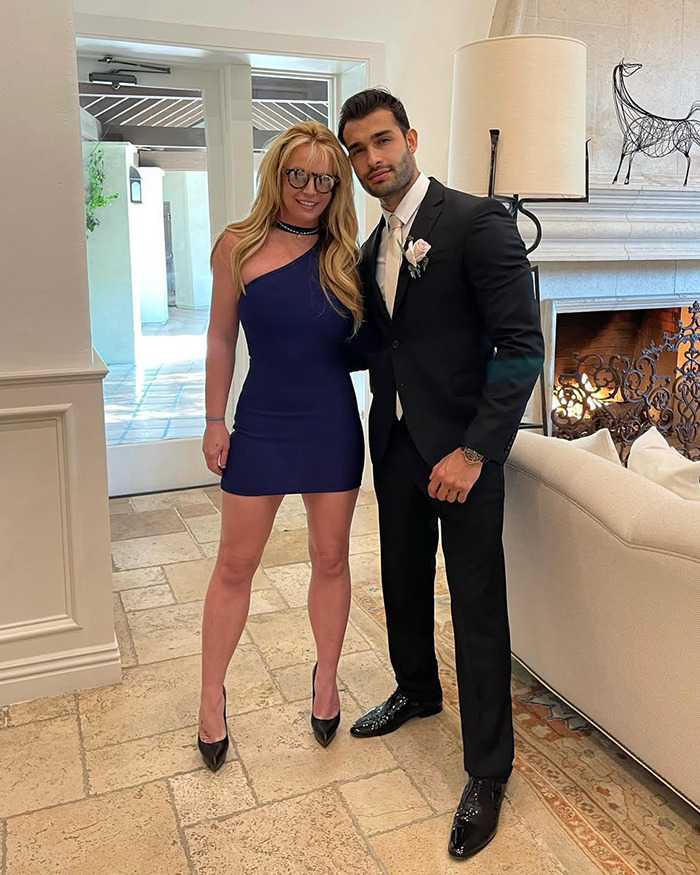 Britney Spears and Sam Asghari dressed formally indoors, posing together on a tiled floor near a fireplace.
